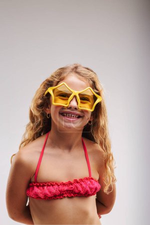 Retrato de media longitud de niña en traje de baño rosa sonriendo a pesar de que no tiene todos sus dientes desarrollados. Lleva gafas en forma de estrella. La vida es todo sobre la alegría.