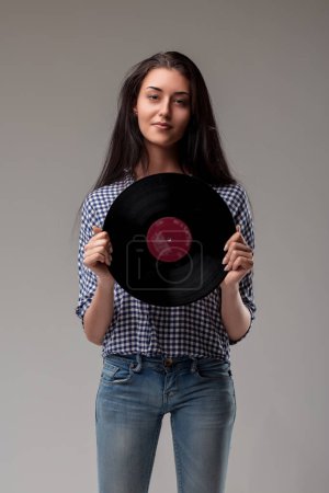 Young woman in blue checked shirt presents a vinyl record, standing firm with a subtle expression