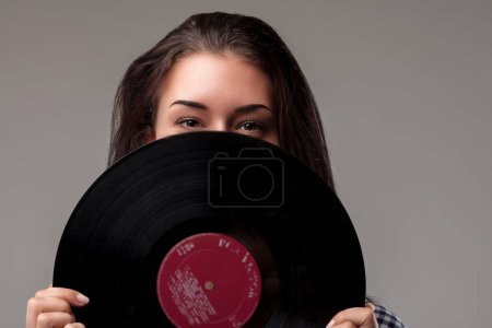 Young woman playfully hides behind a vinyl record, revealing only her eyes, wearing a blue checkered shirt