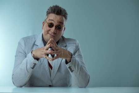 Businessman in grey suit smirks confidently behind desk, fingers interlocked, wearing round sunglasses and displaying various rings