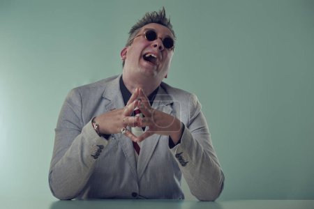 Mad malvado ejecutivo alegre en chaqueta gris y gafas de sol se ríe con estruendo, mostrando una personalidad animada en su escritorio
