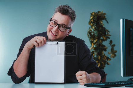 Man in dark shirt and glasses, holding a clipboard with a blank sheet, shows an uncertain and skeptical expression, seated at a desk with a plant and computer, conveying hesitation