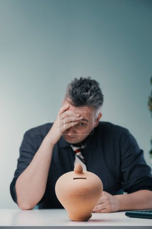 Man in dark shirt and striped tie sits with a frustrated expression, hand on forehead, behind a clay piggy bank on a desk, indicating financial stress and uncertainty about savings