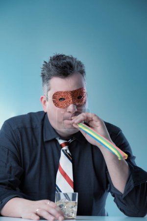 Man in dark shirt and striped tie wears a glittery mask, blowing a party horn while holding a drink, masking his sadness with a forced celebration, portraying deeper emotional pain