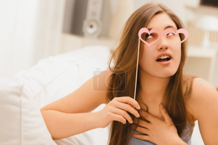 Capturada en un humor juguetón, una mujer mira a través de gafas rosas en forma de corazón, con la mano en su corazón maravillado