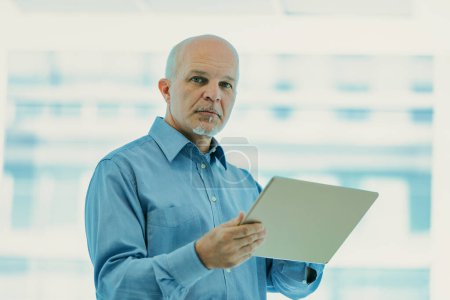 Seasoned professional stands confidently in a well-lit office environment, holding a tablet, portraying determination