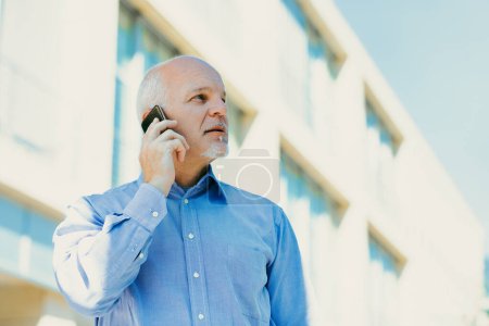 Reifer Mann in blauem Hemd bespricht vor einem modernen Bürogebäude wichtige Angelegenheiten am Telefon und spiegelt Professionalität und Engagement wider