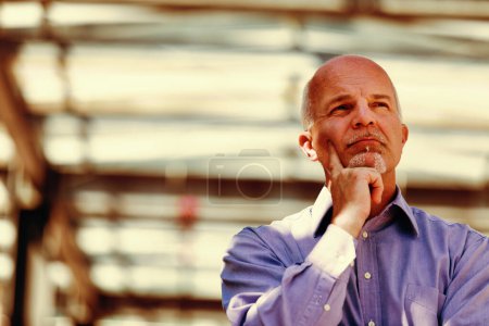 Pensive man in a light blue shirt reflects deeply with his chin rested on his hand, industrial background blurred