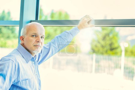 Thoughtful elderly man in blue shirt reflects on his future and family, gazing out of a large window in a sunlit room, contemplating retirement and his grandchildren