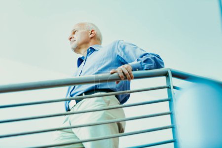 Ein älterer Mann im blauen Hemd lehnt an einem Geländer und blickt nachdenklich in die Ferne, während er über die Vergangenheit und Zukunft des Lebens nachdenkt.
