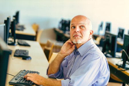In a computer lab, an older man with a grey beard and contemplative gaze looks upward while seated at a workstation, his hand gently resting on his neck, surrounded by empty computer desks