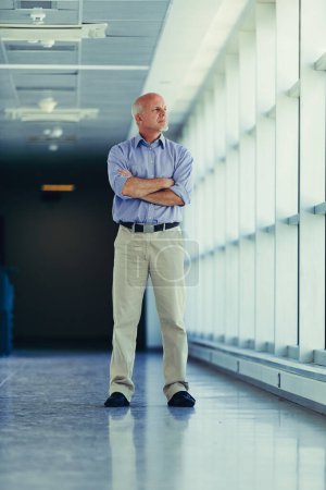 Mature man in business casual attire stands contemplatively in a brightly lit corridor, his arms crossed, reflecting on professional matters