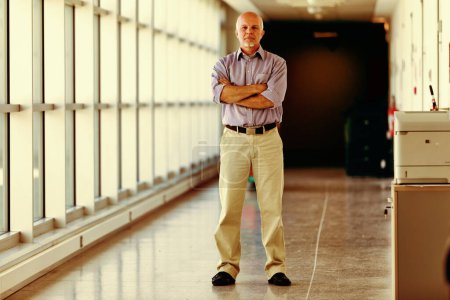 Hombre mayor profesional se detiene en un pasillo junto a grandes ventanales, su postura relajada pero reflexiva, vestido con camisa azul claro