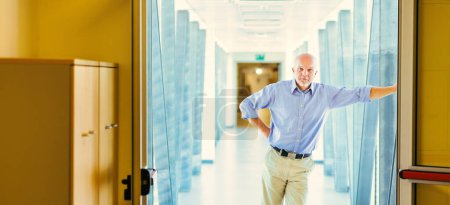 Standing in a bright hallway, an elderly man in a blue shirt leans against a door frame. He looks serious, thinking about future challenges like climate change, job losses, and power concentration