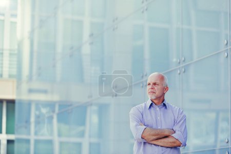 Homme d'affaires mature en chemise bleue pose à l'extérieur d'un complexe de bureaux contemporain, sa posture exsudant autorité et expérience