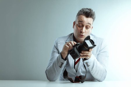Homme d'âge moyen dans un blazer gris clair inspecte une caméra professionnelle avec une expression concentrée, chemise gris foncé en dessous, assis à une surface blanche avec un fond simple