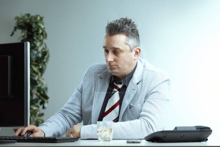 Ein Mann mittleren Alters in hellem Anzug und gestreifter Krawatte arbeitet am Computer, auf dem Schreibtisch liegt ein Glas Whiskey. Sein konzentrierter Ausdruck kontrastiert mit der beiläufigen Anwesenheit von Alkohol bei der Arbeit