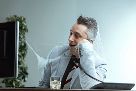 Homme aux cheveux gris pointu, veste de costume gris clair, chemise gris foncé et cravate rayée, couvert de toiles d'araignée, parle au téléphone avec une expression fatiguée, boit sur le bureau, suggérant un très long appel