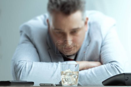 middle-aged man in a light suit stares intently at a glass of whiskey on his desk, battling the urge to drink. His somber expression reflects the internal struggle and the potential consequences
