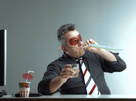 Ein Mann mittleren Alters in dunklem Hemd und gestreifter Krawatte trägt eine rote Glitzermaske und bläst mit einem Whiskeyglas in der Hand in ein Partyhorn. Die festliche Atmosphäre kontrastiert mit seinem Büroumfeld