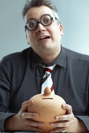 Mann mittleren Alters in dunkelgrauem Hemd und gestreifter Krawatte, mit spitzen grauen Haaren und übergroßer runder Brille, unbeholfen lächelnd, während er ein Keramik-Sparschwein in der Hand hält, das schlechtes Finanzmanagement symbolisiert, schlicht 
