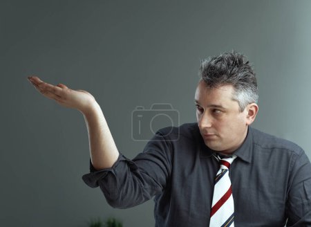 Mann in dunklem Hemd und gestreifter Krawatte hebt die Hand nach oben und bietet oben Platz für eine Produktpräsentation. Sein konzentrierter Blick legt die Bereitschaft nahe, das Objekt zur Schau zu stellen