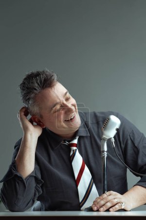 Wearing a dark shirt and striped tie, the man smiles and scratches his head while speaking into a vintage microphone. His cheerful expression and relaxed posture indicate a fun podcast episode
