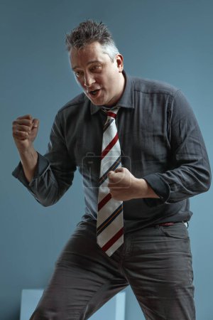 middle-aged man in a dark shirt and striped tie clenches his fists, expressing intense determination. His short, spiky hair adds to his dynamic, focused demeanor against a plain background