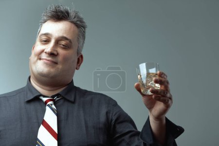 man, dressed in a dark shirt and striped tie, raises a glass of whiskey. His salt-and-pepper hair and slight smile suggest a relaxed, contented mood in a casual setting
