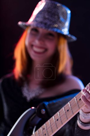 Junge Frau mit kastanienbraunem Haar, glitzerndem Hut und dunklem Glitzertop, spielt E-Gitarre. Ihr Lächeln und ihre selbstbewusste Pose auf der Bühne vermitteln ihre Begeisterung für die darstellende Kunst