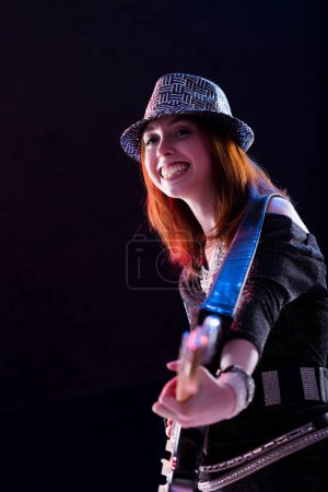 femme aux cheveux auburn, vêtue d'un chapeau pailleté, haut sombre, et un jean, sourit tout en jouant une guitare électrique. Son comportement confiant et joyeux souligne sa passion pour la musique