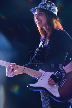 junge Frau mit roten Haaren, gemustertem Hut und schwarzem Glitzeroutfit, spielt unter Bühnenlicht eine E-Gitarre, lächelt selbstbewusst, mit bunten Lichtreflexen um sich herum