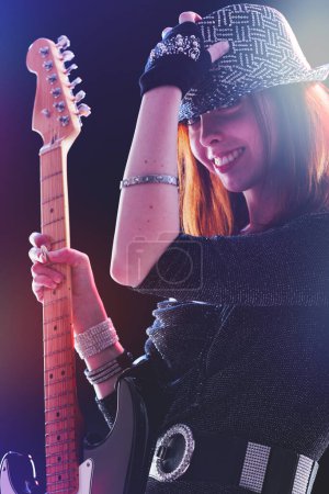 Under vibrant stage lights, a smiling young woman with red hair and a sequined hat strums her electric guitar passionately, her fingers poised expertly on the strings