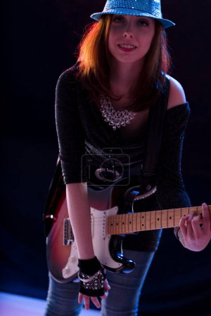 Auburn-haired woman in a sparkling hat and dark glittery top smiles while playing an electric guitar on stage, radiating confidence and joy in her musical performance