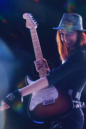femme aux cheveux roux, vêtue d'une tenue noire chatoyante et d'un chapeau élégant, joue une guitare électrique sur scène, son sourire lumineux et contagieux sous les lumières vives