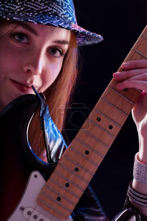 young woman with auburn hair, dressed in a glittery hat and dark top, plays an electric guitar with a smile. Her joyful performance highlights her passion and confidence