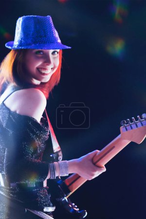 Enthusiastisch spielt sie ihre E-Gitarre, eine junge Frau mit roten Haaren und Paillettenhut leuchtet unter dem Bühnenlicht, ihr schwarzes Outfit schimmert bei jeder Bewegung