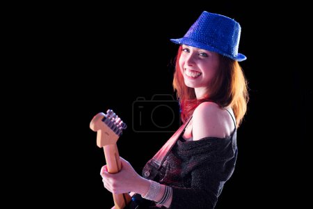 Frau mit kastanienbraunem Haar und glitzerndem Hut spielt lächelnd eine E-Gitarre. In dunklem Top und Jeans strahlt sie auf der Bühne Zuversicht und Freude aus