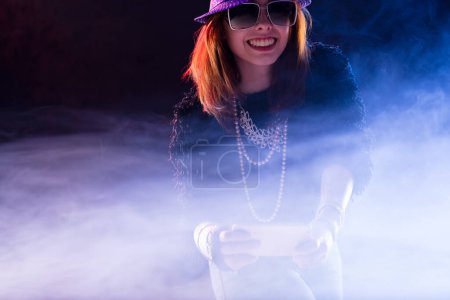 young woman, with auburn hair and wearing a sequined purple hat and sunglasses, uses her smartphone. Surrounded by smoke, she epitomizes immersion in virtual worlds through telematics and the internet