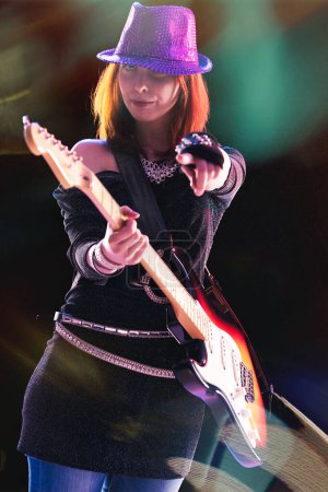  freudige junge Frau mit roten Haaren, in schwarzem Glitzerensemble und Paillettenhut, spielt gekonnt ihre E-Gitarre, Bühnenlichter werfen bunte Spiegelungen um sich