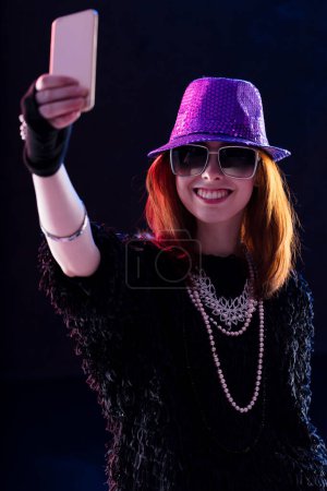 Auburn-haired woman in a purple sequined hat and sunglasses holds a smartphone, standing in a smoky environment. Her attire and engagement with the device illustrate being absorbed in an online scene