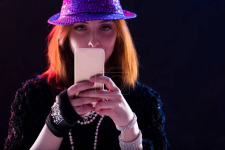 Woman with auburn hair, a purple hat, and sunglasses holds a smartphone, surrounded by smoke. Her focus on the phone highlights how telematics and the internet can immerse one in a virtual scene