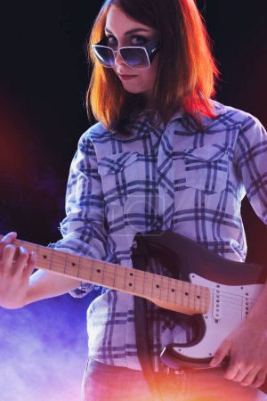 young woman with red hair, wearing a plaid shirt and sunglasses on her head, plays an electric guitar with a wide, enthusiastic smile, surrounded by colorful stage lights