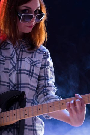 mujer joven con pelo castaño, con camisa a cuadros y gafas de sol, toca una guitarra eléctrica con una sonrisa brillante. Su postura enérgica resalta su entusiasmo y confianza en la realización