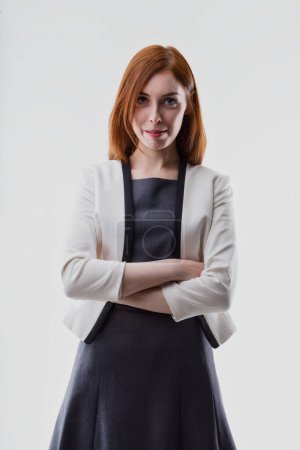 Woman with auburn hair, wearing a white blazer over a dark dress, crosses her arms with confidence. Her professional appearance suggests a role in business or leadership