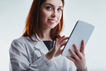 With a gentle smile, a red-haired family doctor in a white lab coat uses a tablet, emphasizing her commitment to compassionate care and modern medical technology