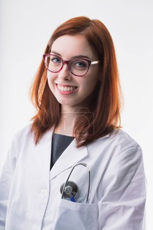 Smiling young woman with auburn hair wears glasses and a white lab coat, with a stethoscope in her pocket. Her friendly demeanor and professional appearance convey competence and approachability