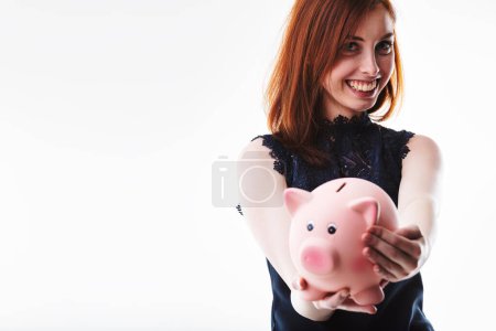 Smiling woman with auburn hair in a dark lace top holds a pink piggy bank forward. Her happy expression highlights the positive aspect of saving money and financial security