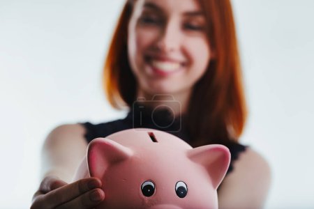 Smiling broadly, a red-haired woman in a black lace top holds a pink piggy bank in both hands, her cheerful demeanor indicating happiness and a sense of financial security