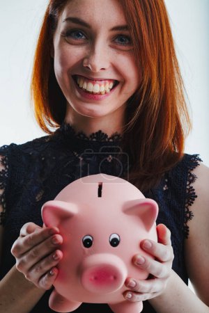 With a wide smile, a red-haired woman in a black lace top displays a pink piggy bank, her expression conveying joy and the importance of saving money